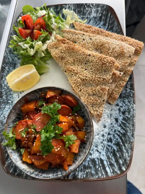 A bowl of spiced sausage and potato cooked down in tomato, beside triangles of warm sesame bread and a dressed salad.